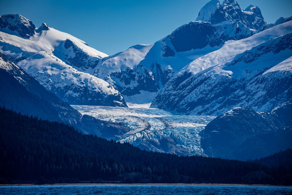 Massive blue glacier flows between snow-covered mountain peaks toward an alpine lake.