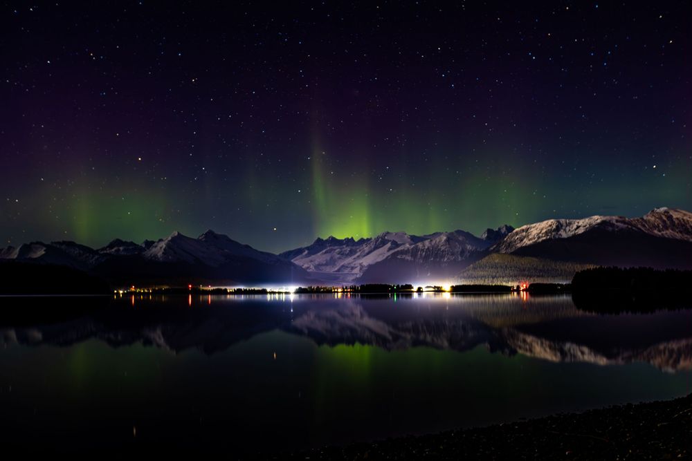 Northern lights dance above snow-covered mountains reflected in still lake waters with distant town lights.