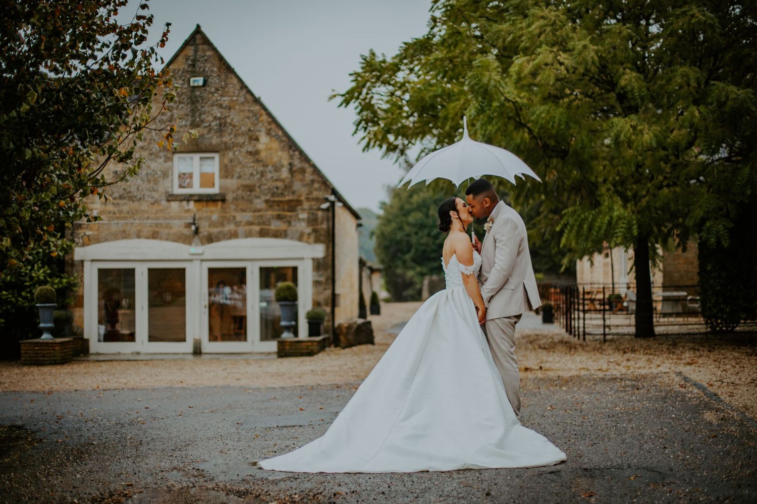 Bride and groom kissing under an umbrella in the rain at Lapstone Barn Wedding Venue, The Cotswolds - RELM Photography