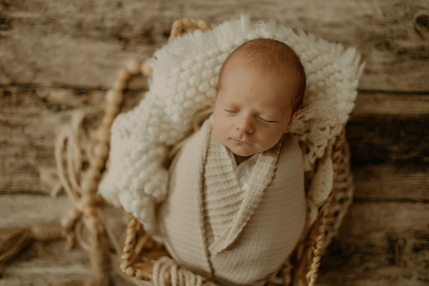 Newborn baby sleeping peacefully wrapped in neutral knit blanket against wooden backdrop.