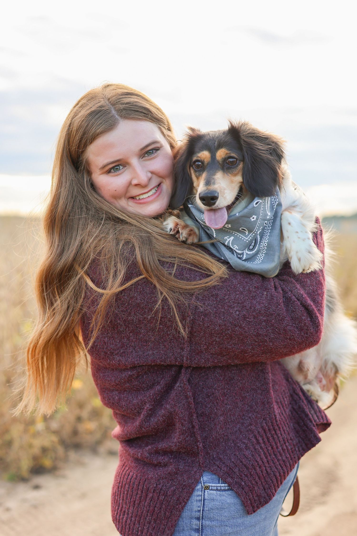 An adorable mixed breed dog wearing a blue bandana is held lovingly while outdoors at sunset.