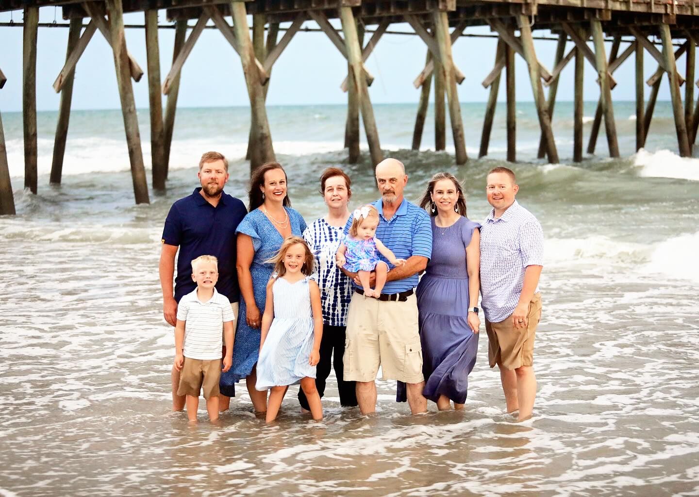 A group poses together under a wooden pier at the beach during sunset.