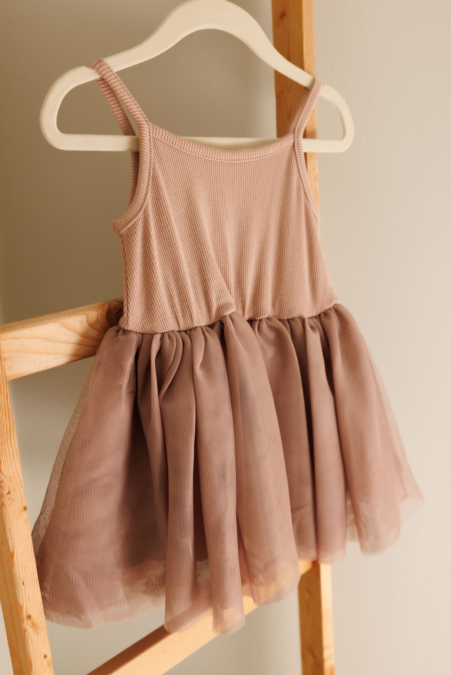Pink ballet-inspired dress with ribbed tank top bodice and tulle skirt hanging on white hanger against wooden ladder.