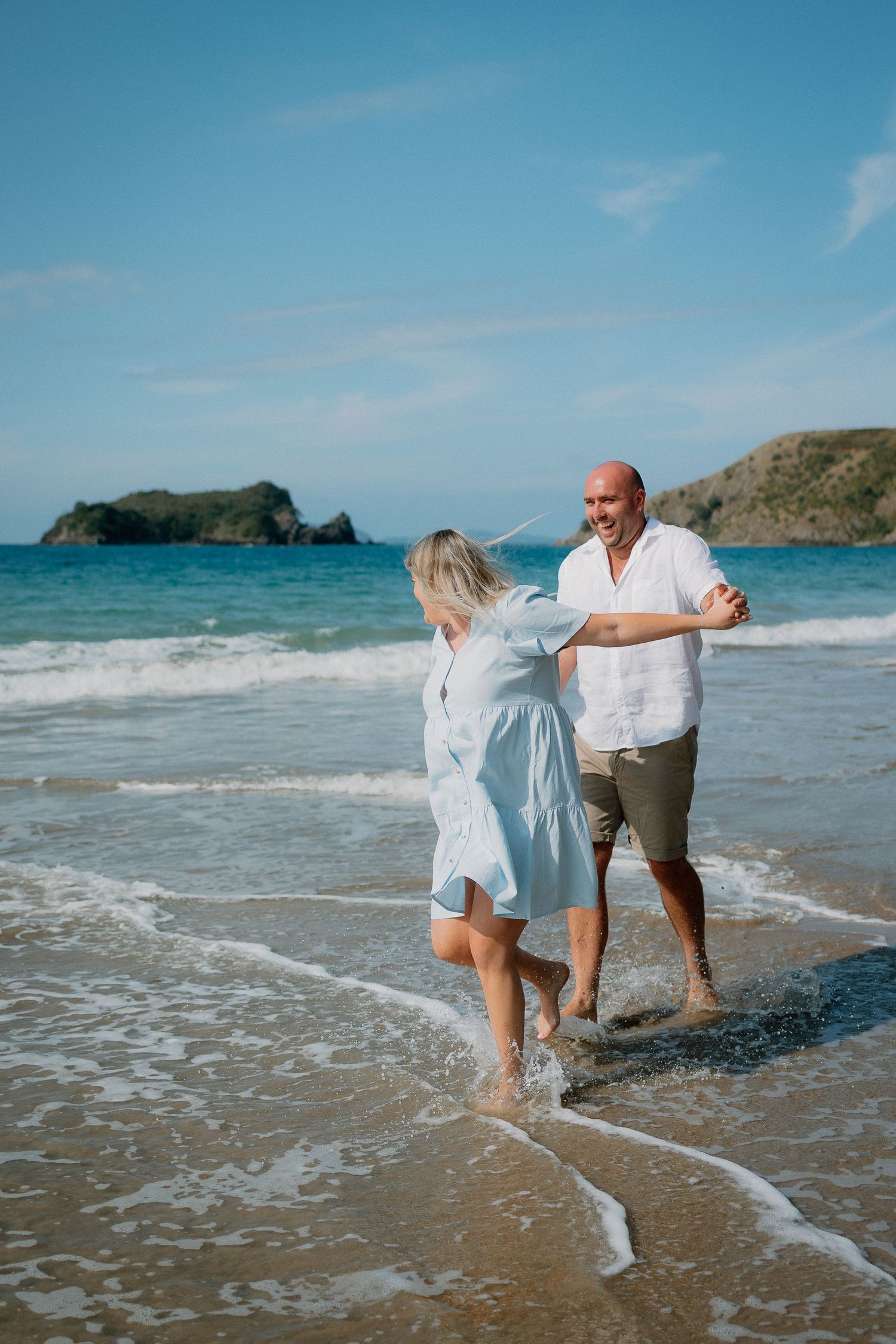 A couple walks together along a tropical beach with turquoise waters and rocky islands in the background.