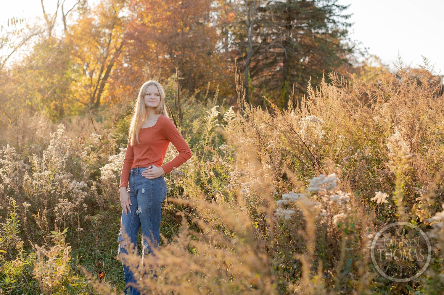 Outdoor portrait with orange sweater amid golden autumn grasses and fall foliage.