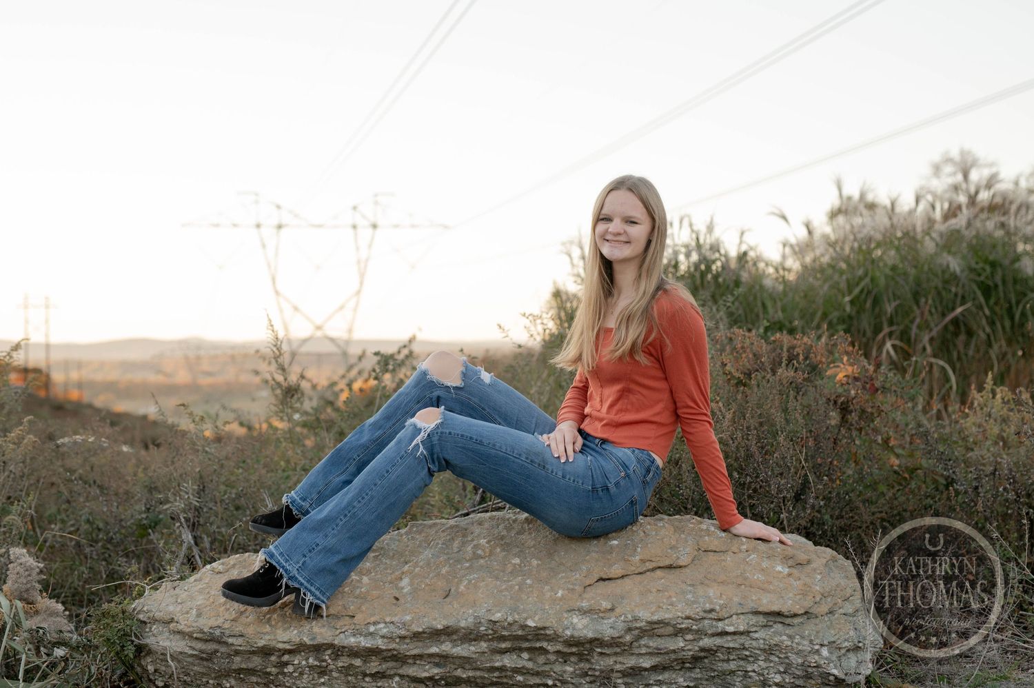A person in orange cardigan and jeans sits on a rock during sunset with power lines in background.