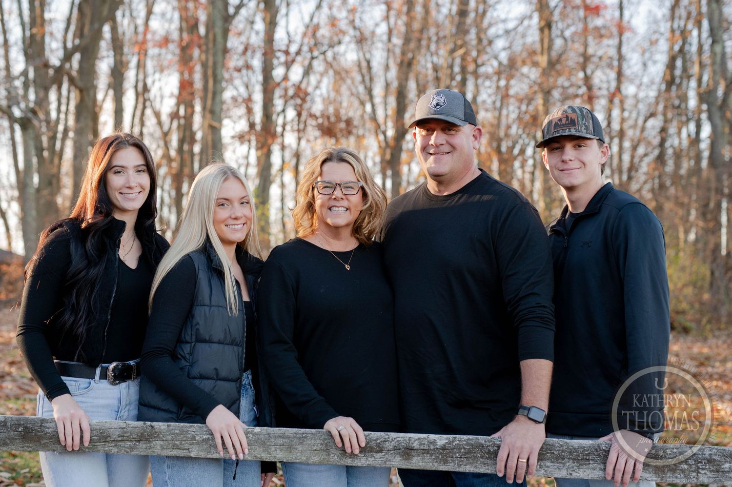 A group of five people wearing dark clothing pose along a wooden fence in an autumn forest setting.