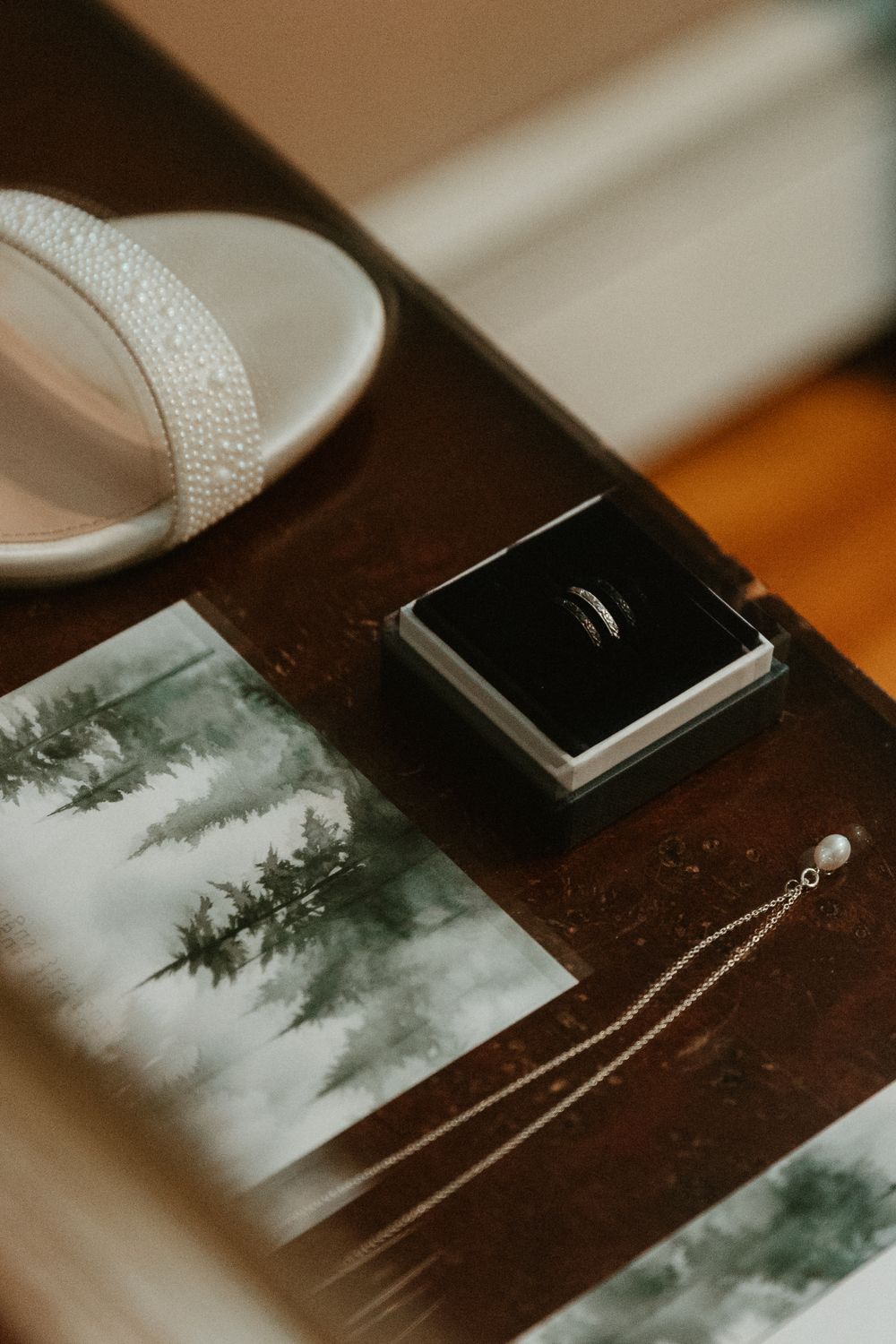 Wedding details including a ring box, white shoe, and forest-themed stationery on a dark wooden surface.