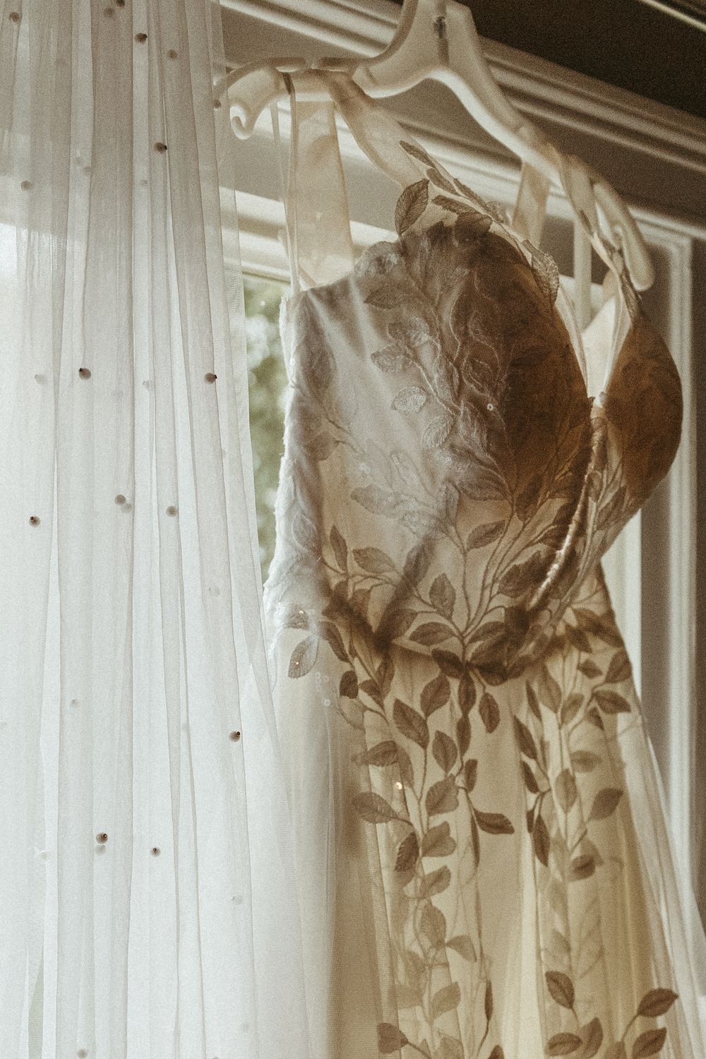 White lace wedding dress hanging against sheer curtains in natural window light.