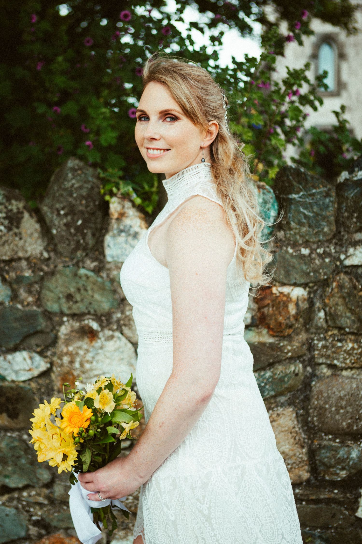 A person in an elegant white lace wedding dress holds a vibrant yellow and orange floral bouquet beside a stone wall.