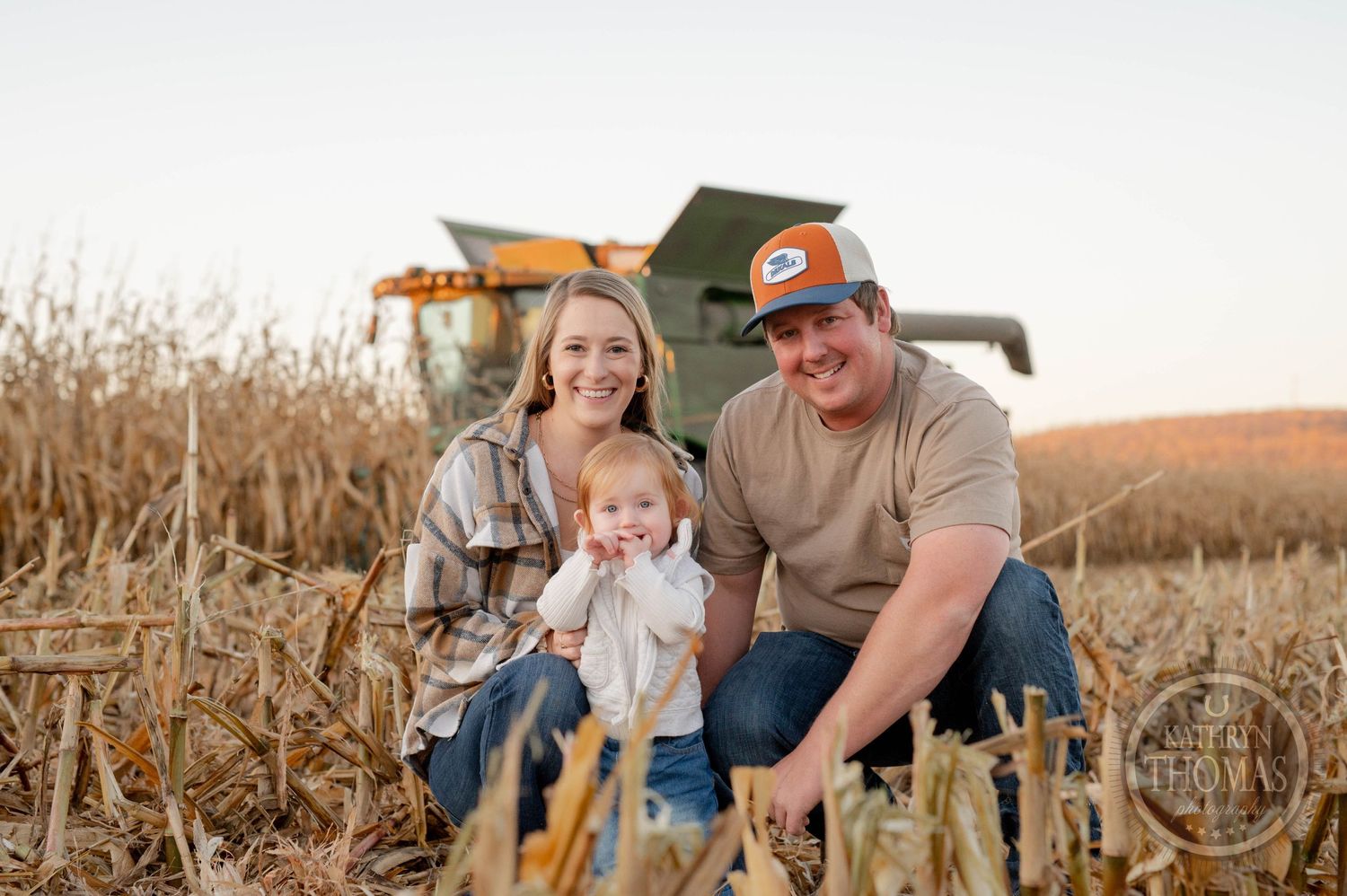 Family portrait in autumn cornfield with John Deere equipment during harvest season.