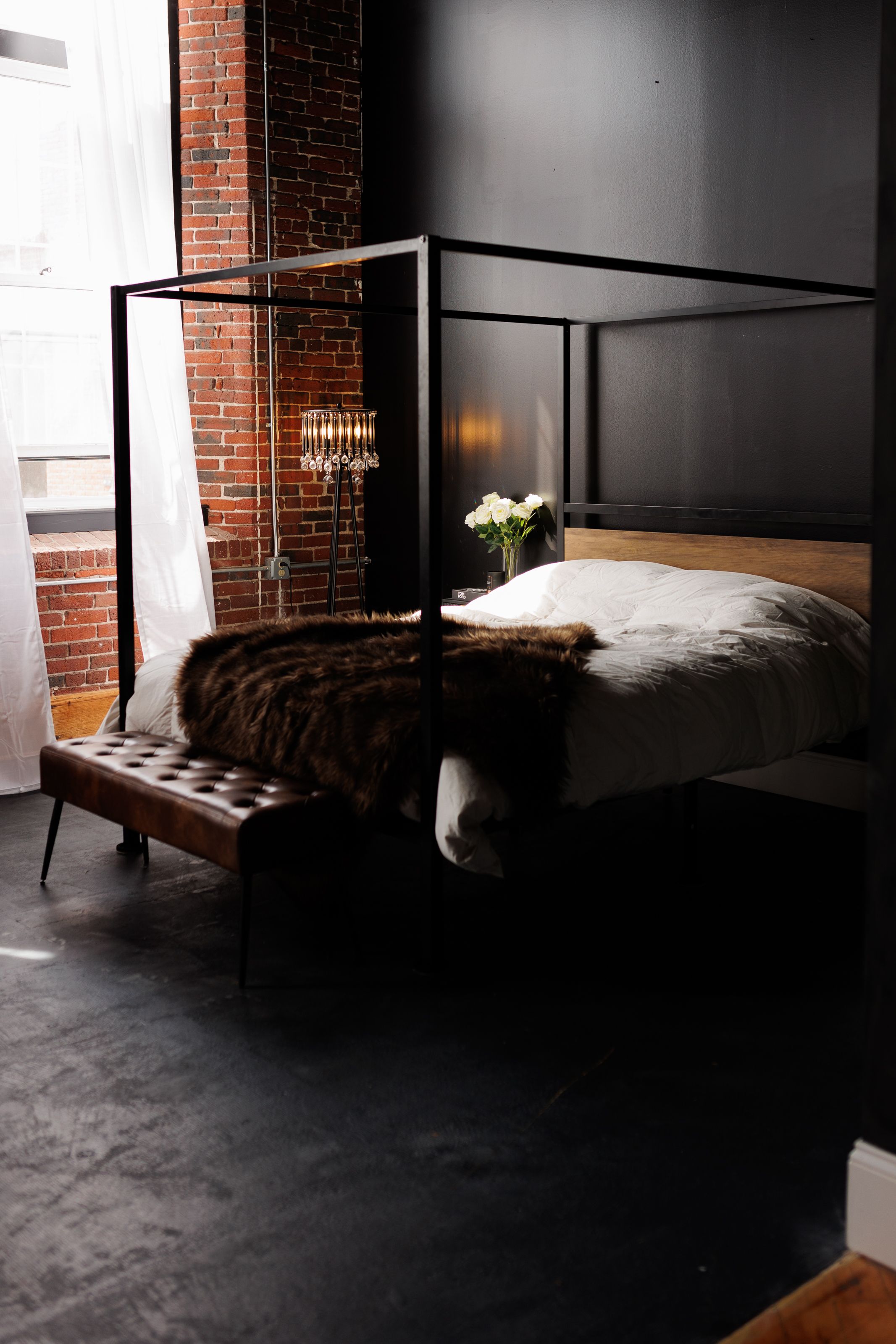Modern industrial bedroom with black canopy bed, leather bench, and exposed brick wall illuminated by warm lighting.
