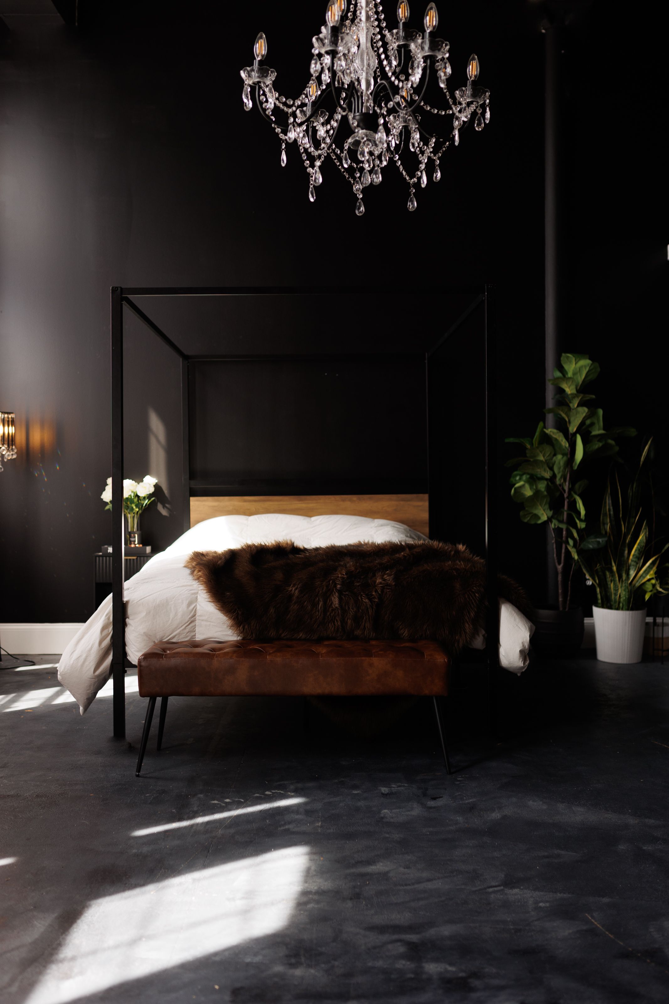 A modern bedroom with black walls features a crystal chandelier, canopy bed, and brown fur throw in dramatic lighting.