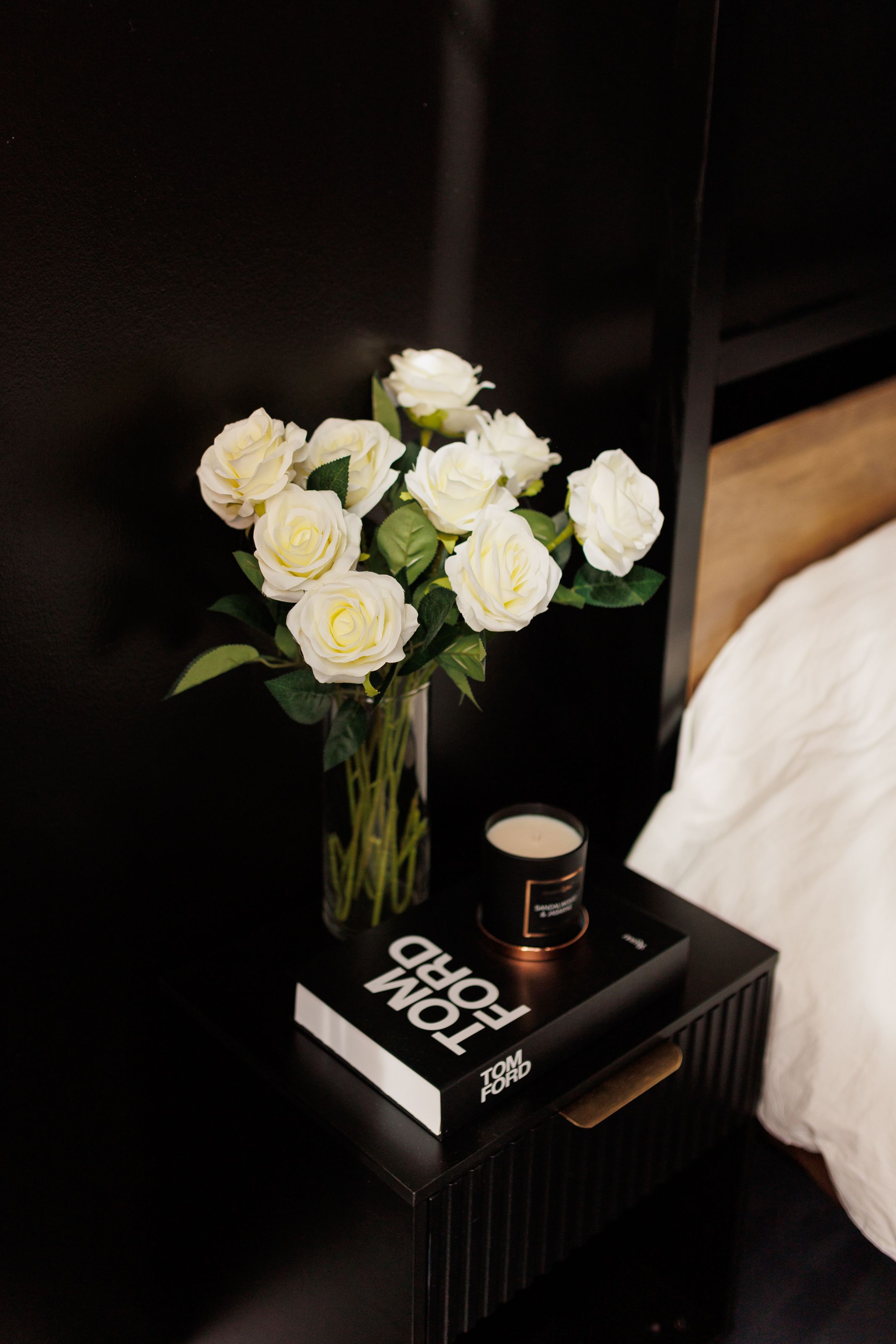 White roses in a glass vase sit atop a dark bedside table with a black book and candle against a dark background.