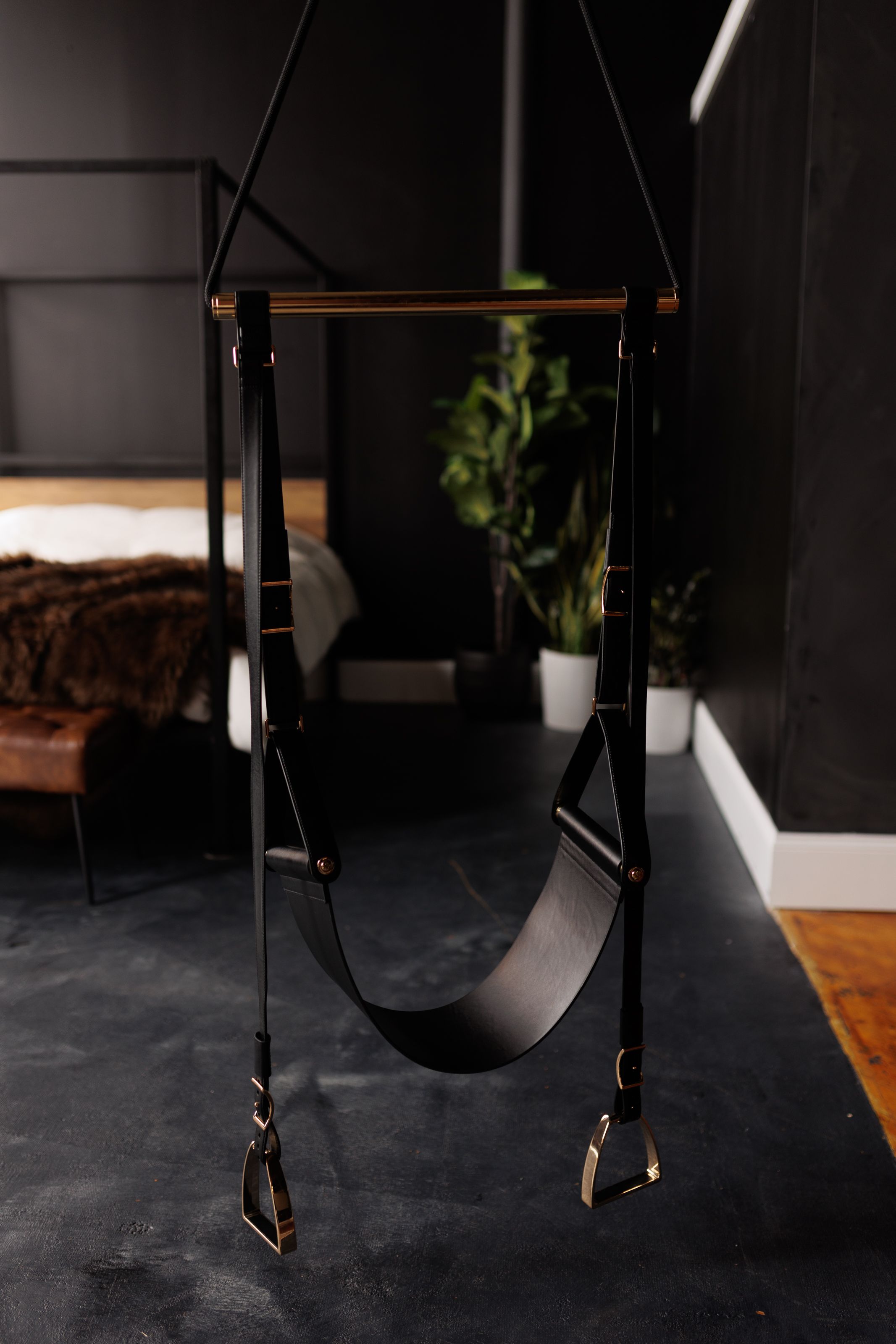 Modern leather swing with wood beam and black leather straps mounted against dark wall in minimalist interior space.