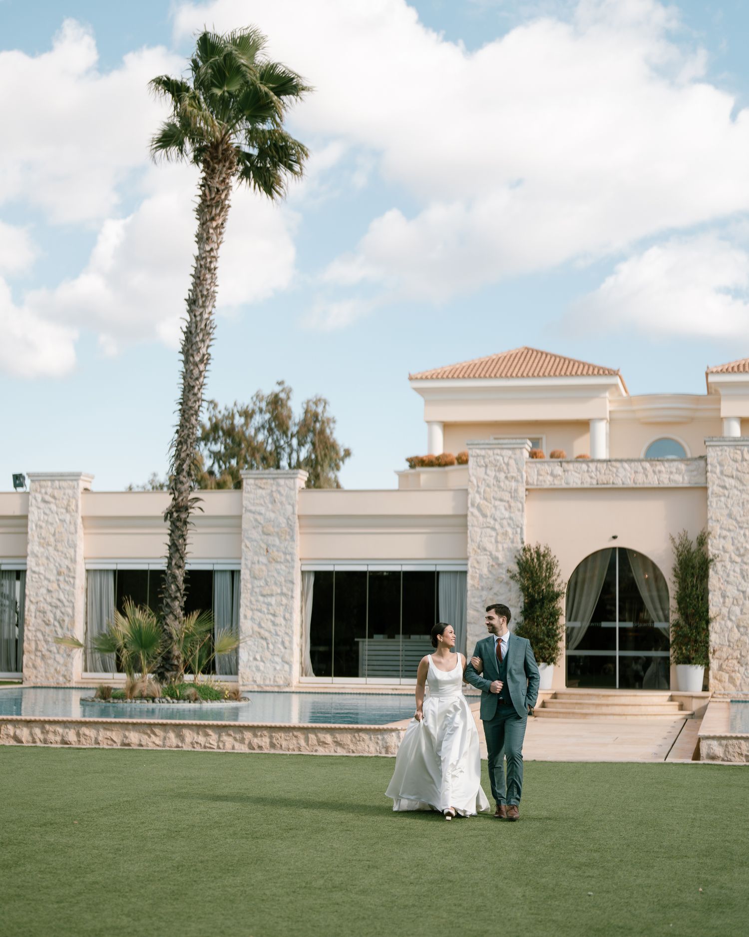 A wedding couple poses on a lawn with a palm tree and elegant white building behind them. next day wedding photoshoot.