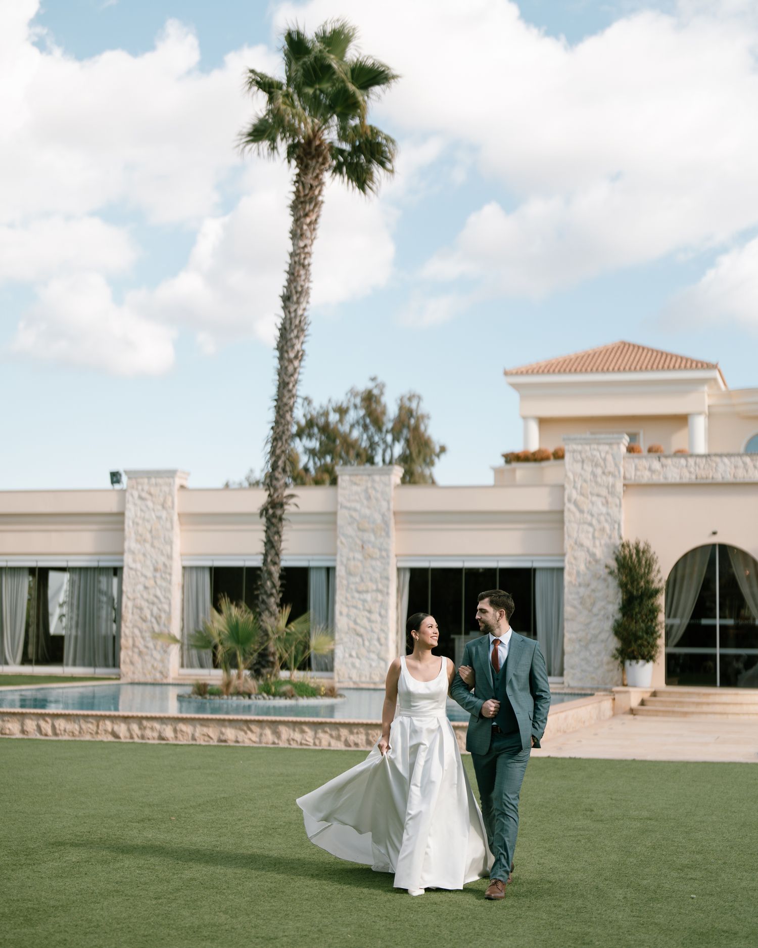 A couple walks together on manicured grounds with a Mediterranean-style building and palm tree in the background. next day wedding.