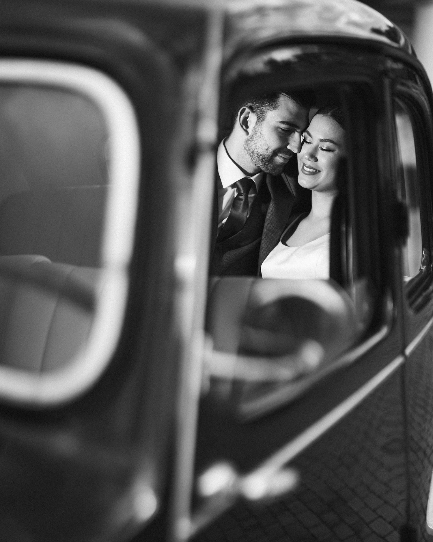 A black and white photo of newlyweds sharing an intimate moment in a vintage car.