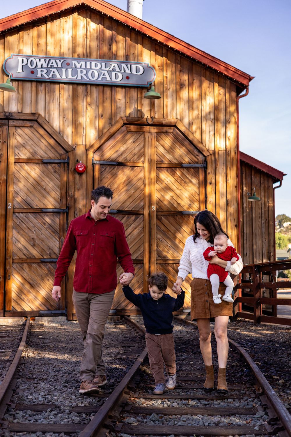 Family walking along train tracks at Old Poway Park, mom holding baby girl, both parents holding little boy’s hands during holiday session.