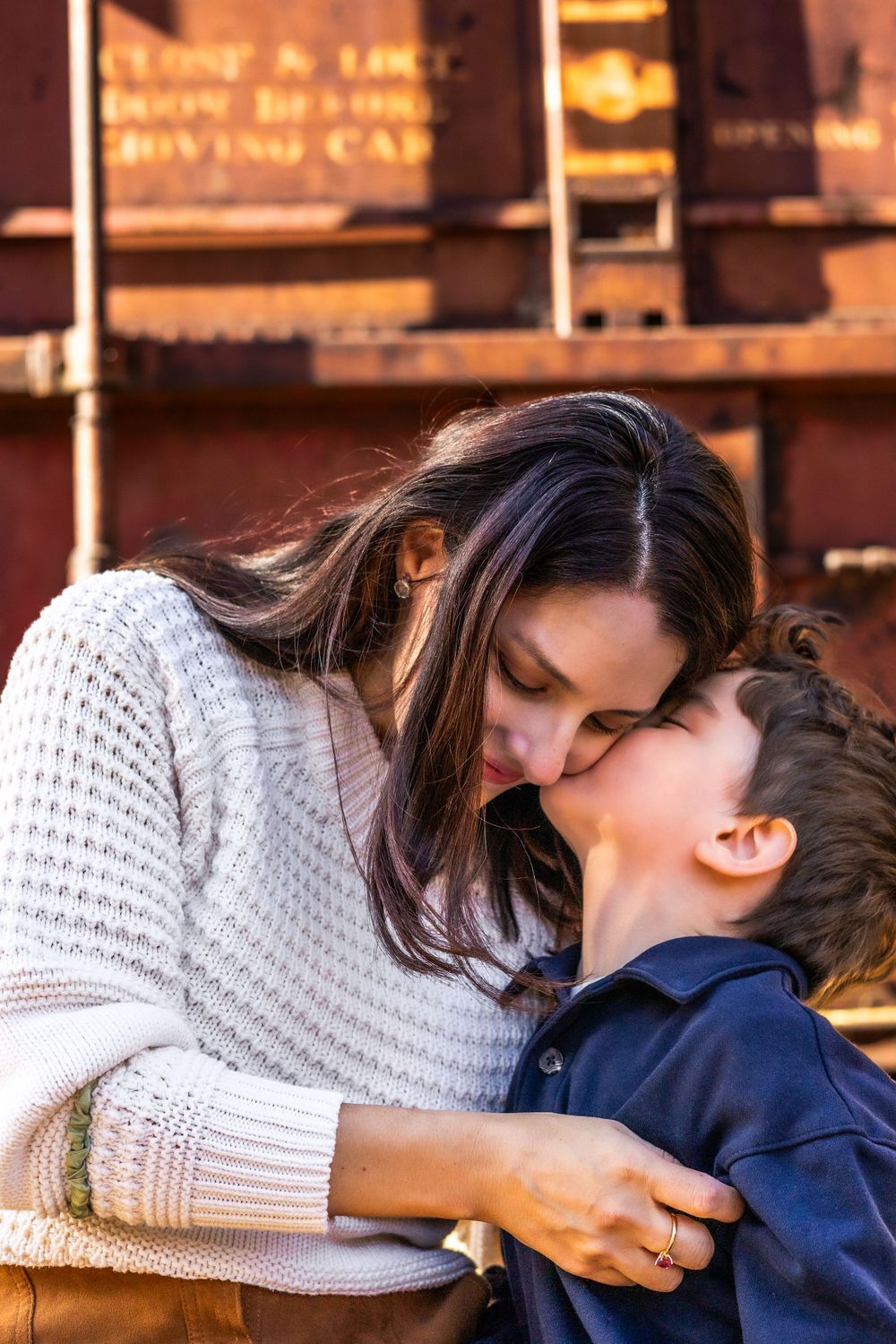 Close-up of little boy kissing mom's cheek as she looks down and smiles with her eyes at Old Poway Park in a precious motherhood moment.