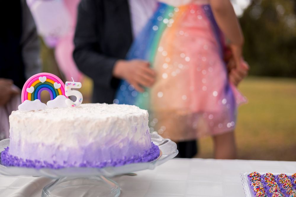 Close-up of birthday cake with rainbow topper and number 5, with little girl’s sparkly, colorful dress softly blurred in warm light.