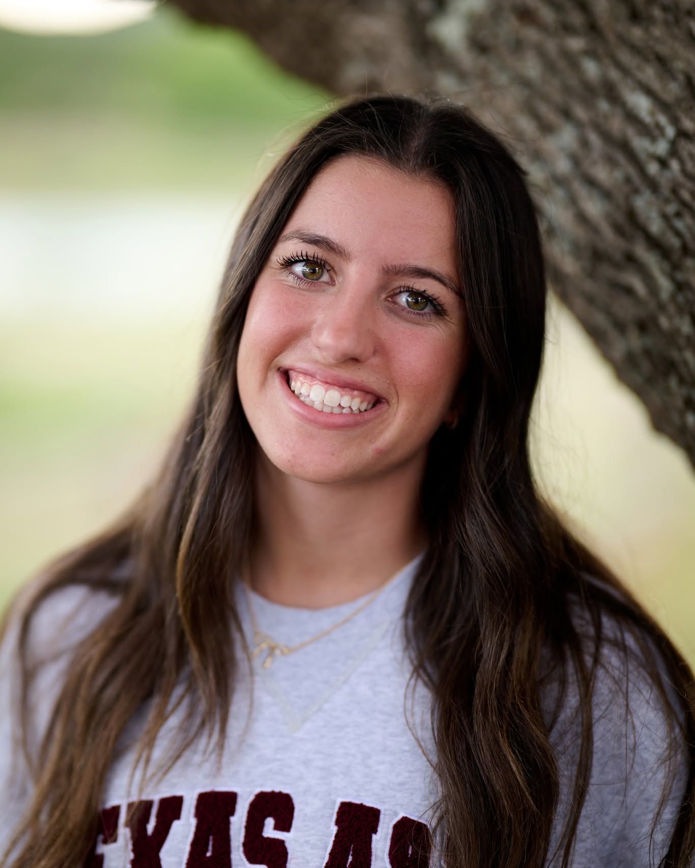 A portrait photo shows a smiling person with long dark hair wearing a light gray Texas A&M shirt while standing by a tree.