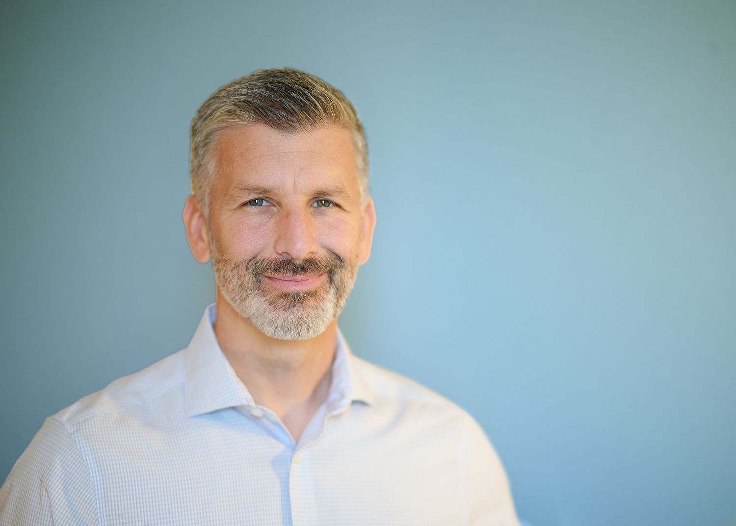Professional headshot of a middle-aged person in a light blue button-down shirt against a blue background.