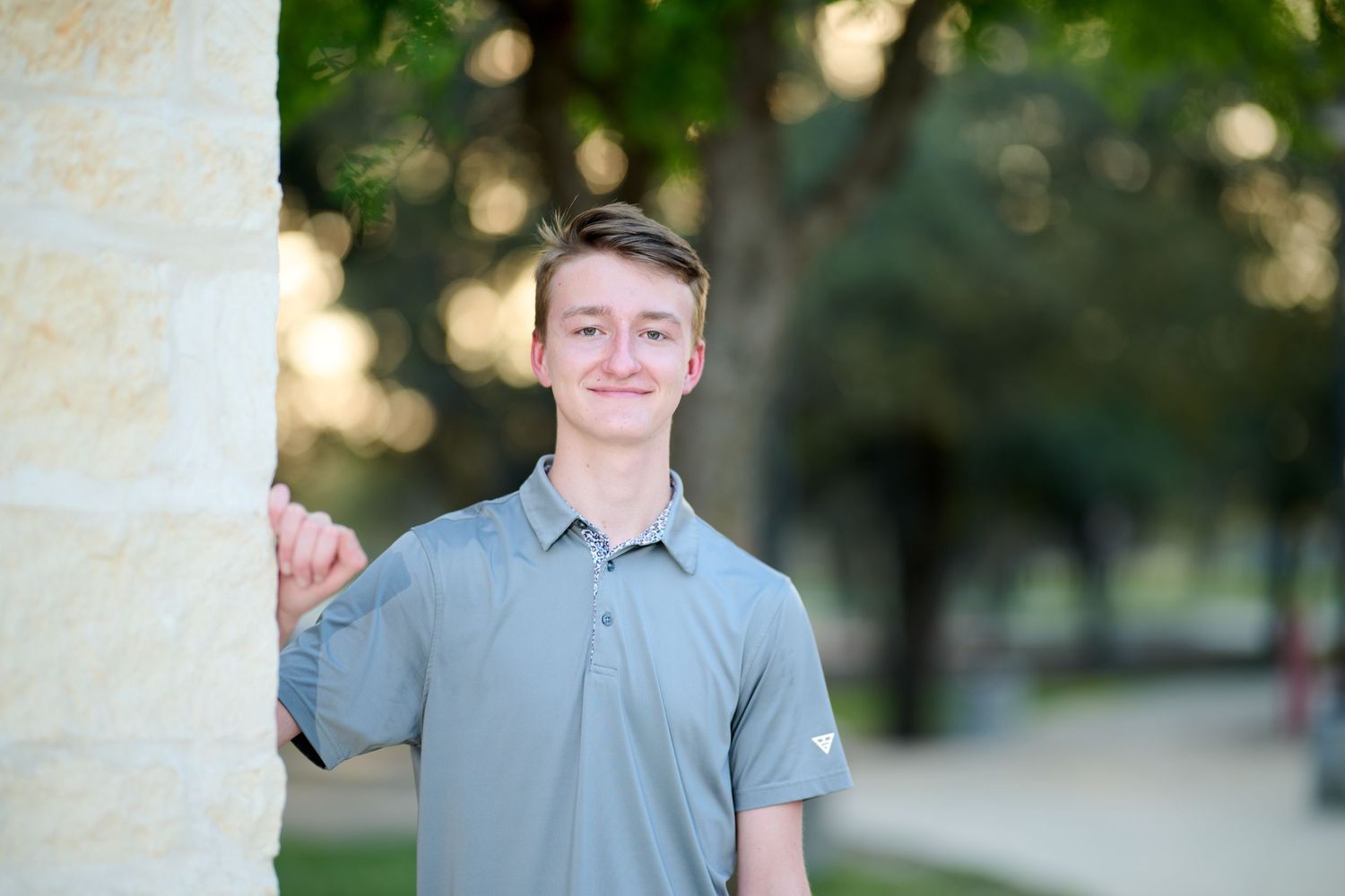 A young man in a gray polo shirt stands casually next to a stone column outdoors.