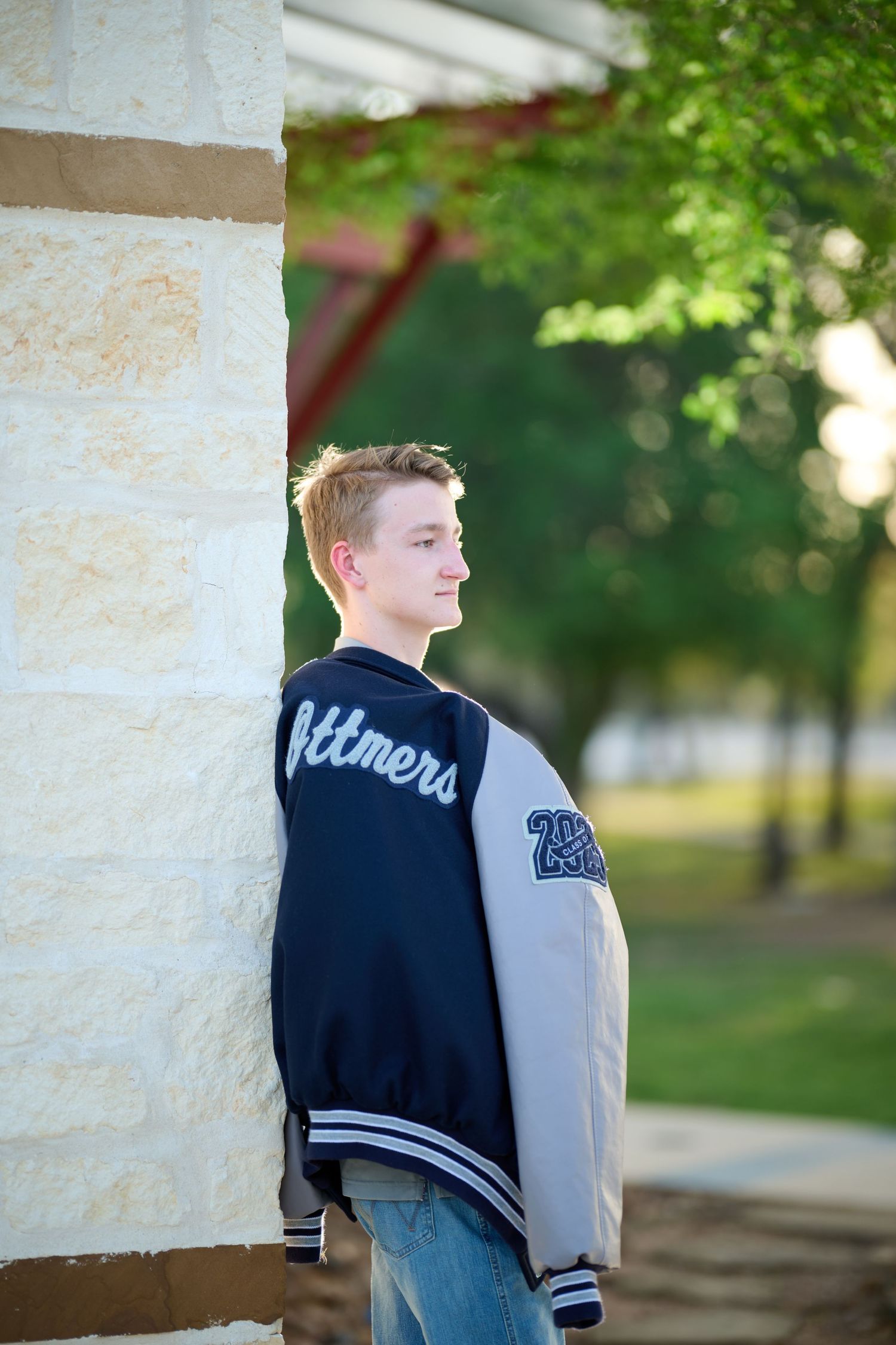 Student wearing a varsity letterman jacket poses against a white stone column outdoors.