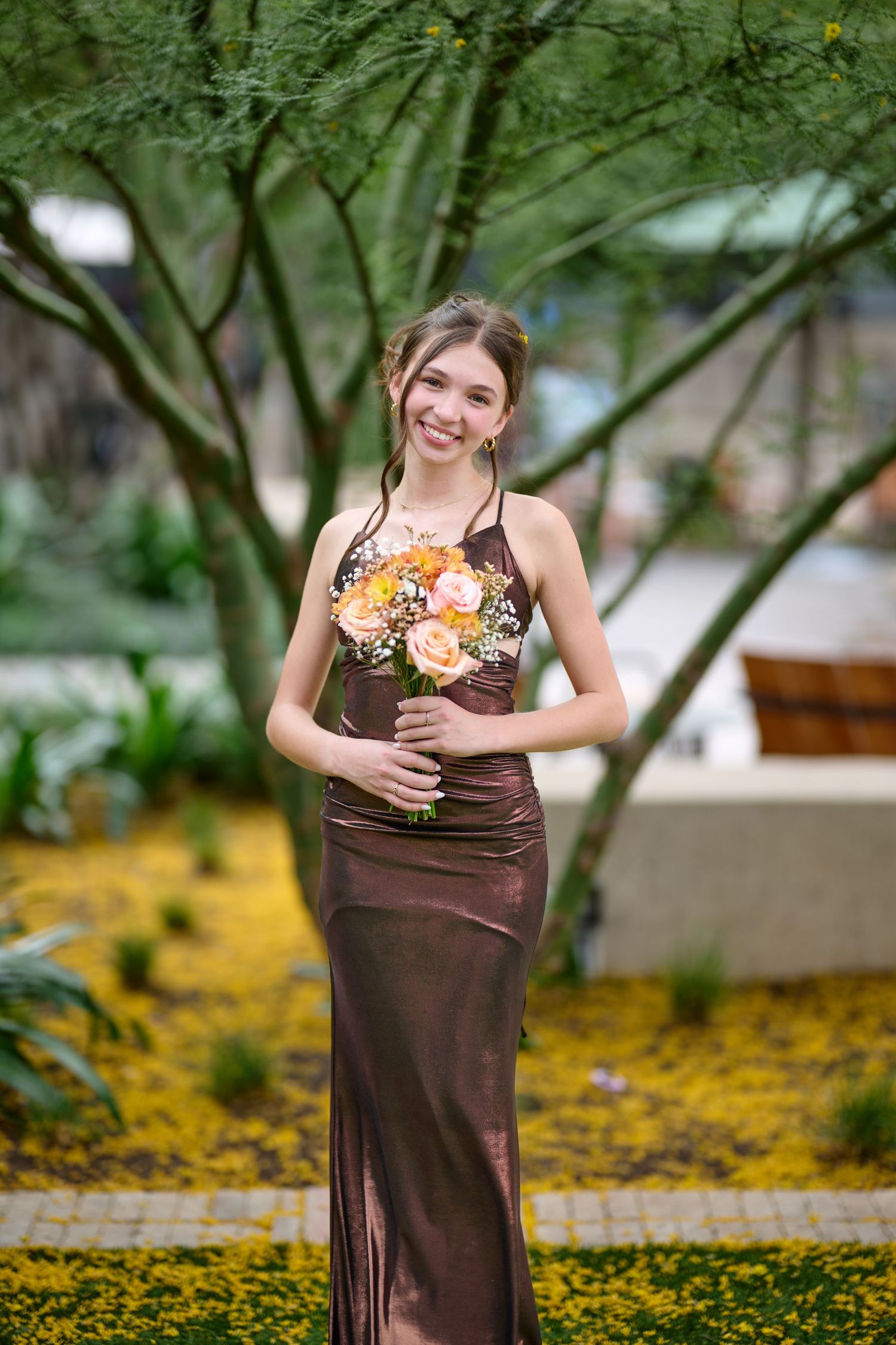 Person in a brown formal dress holds a bouquet of flowers while posing in a garden setting.