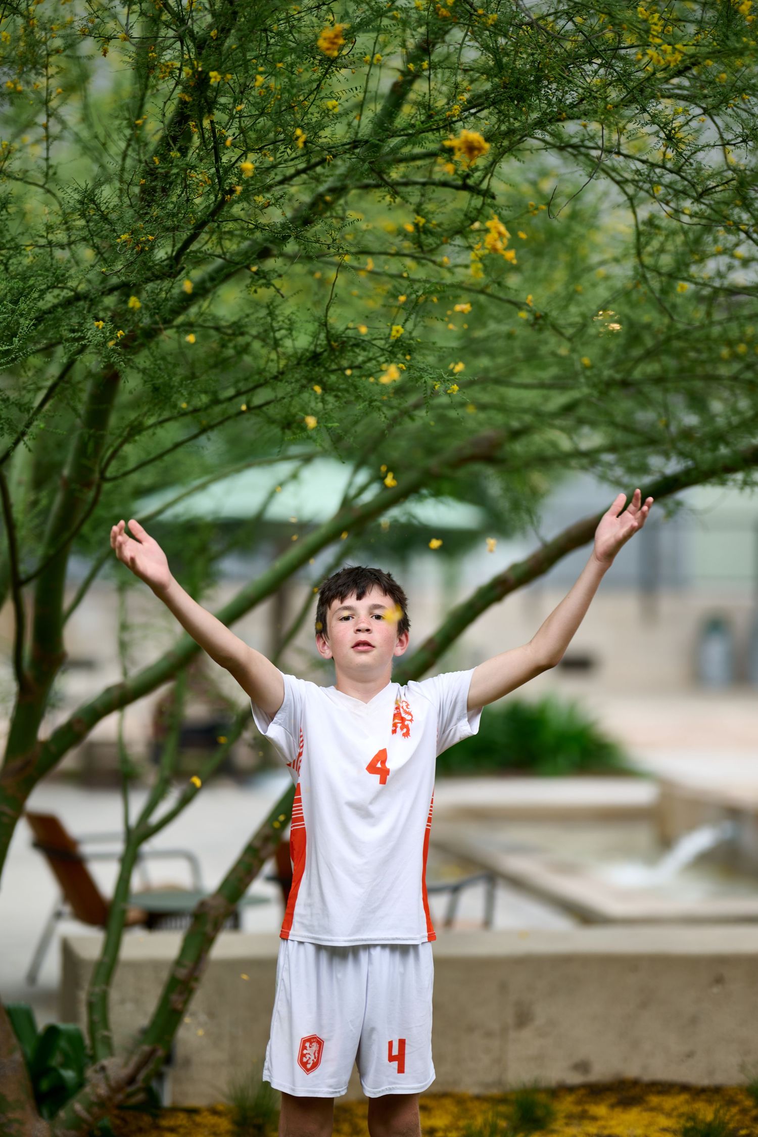 Young soccer player in white uniform celebrates with raised arms under a flowering tree.