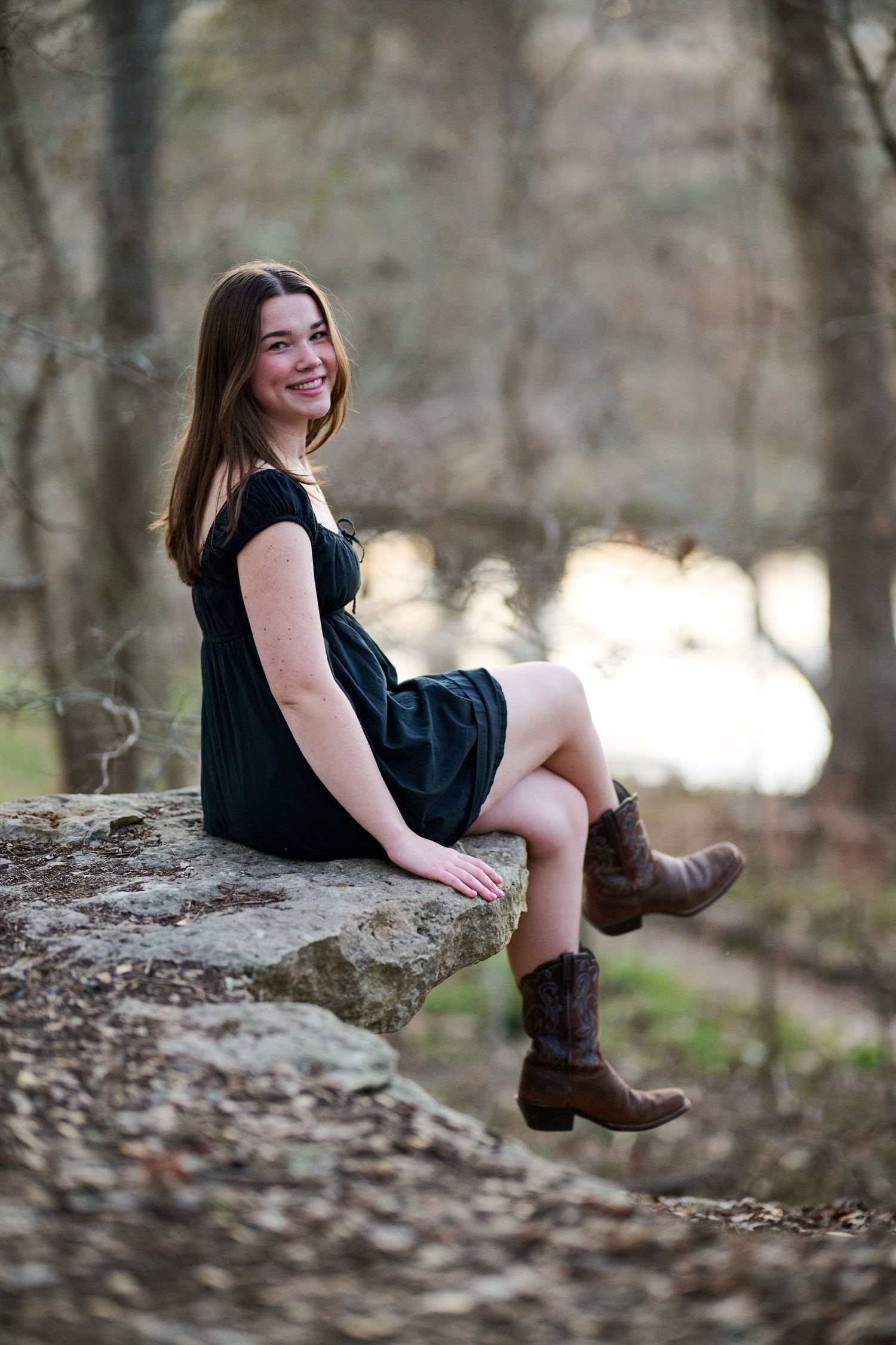 Person in a black dress and boots sits casually on a rock in a wooded setting.