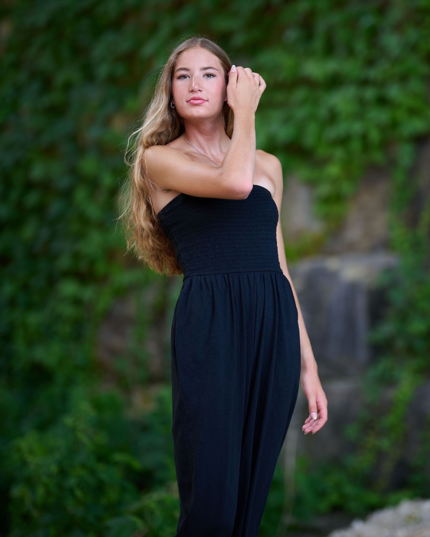 A young woman in a black dress poses gracefully outdoors with a wooded green background.