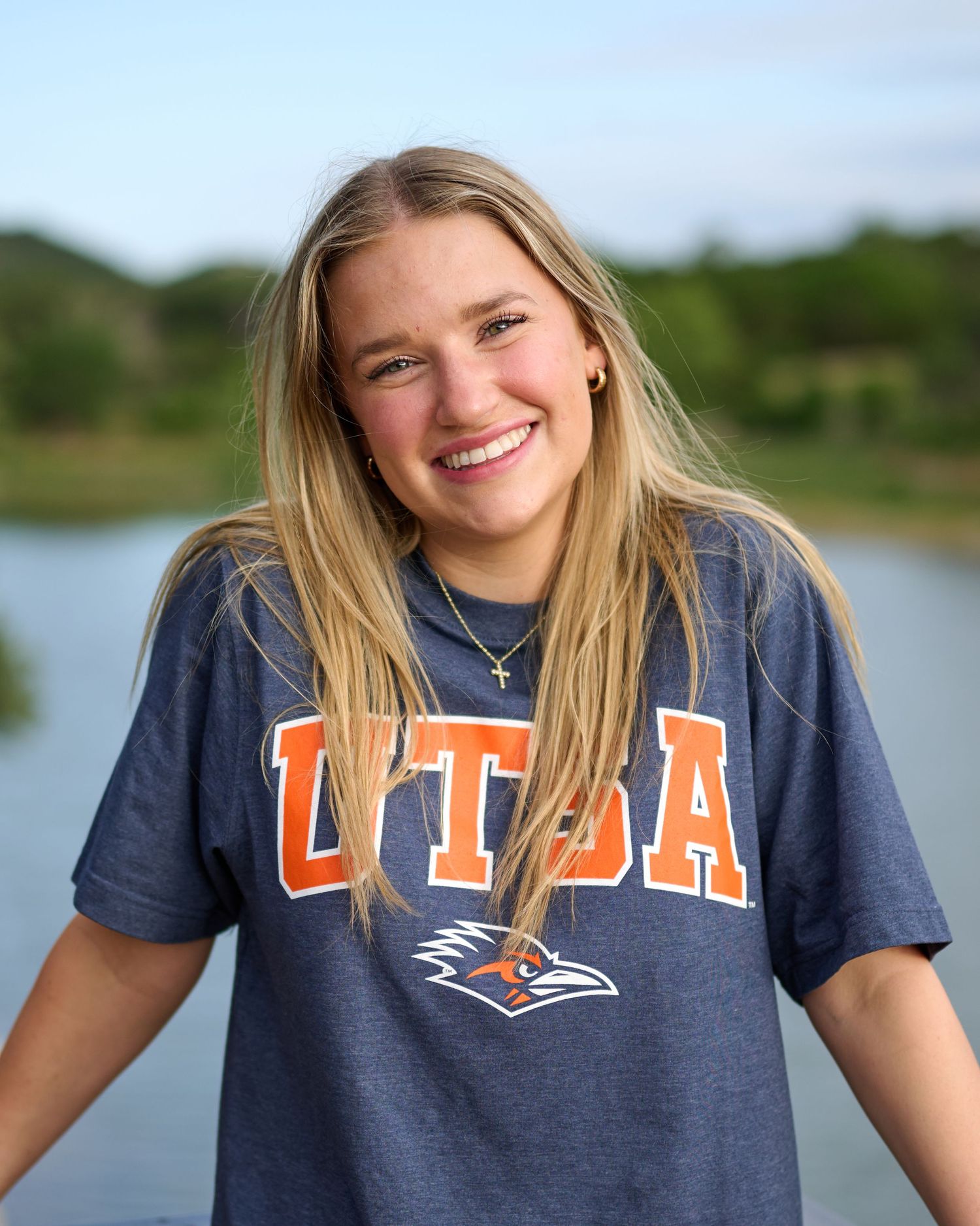 Person wearing UTSA college t-shirt smiling outdoors near water.