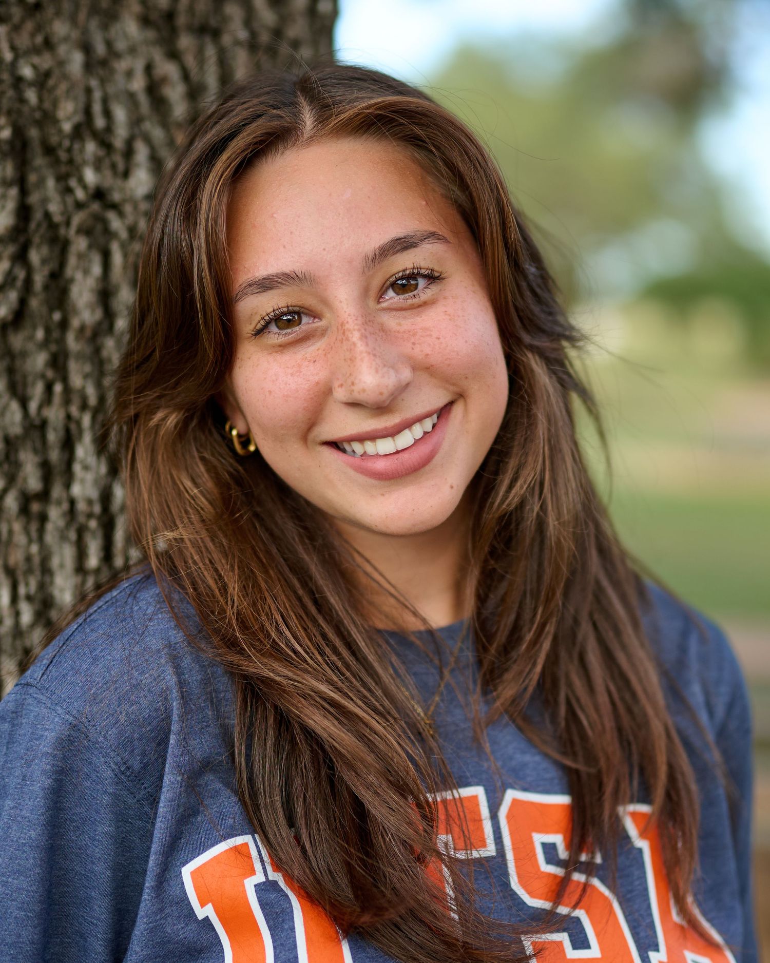 Student wearing a USA college sweatshirt smiles while leaning against a tree trunk outdoors.