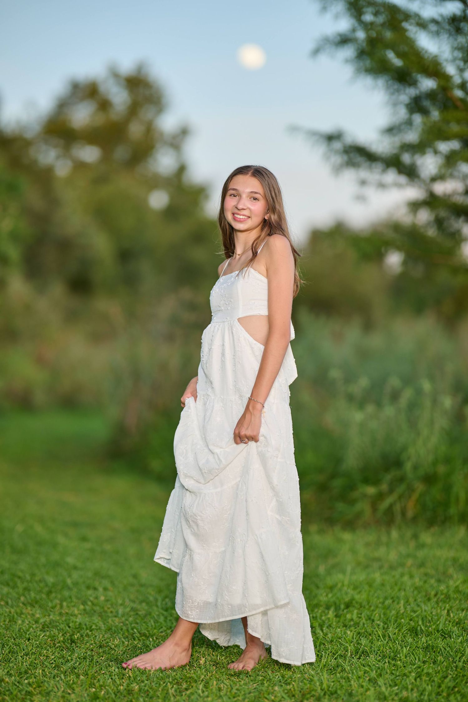 Outdoor portrait in flowing white dress standing barefoot in grassy field at dusk.