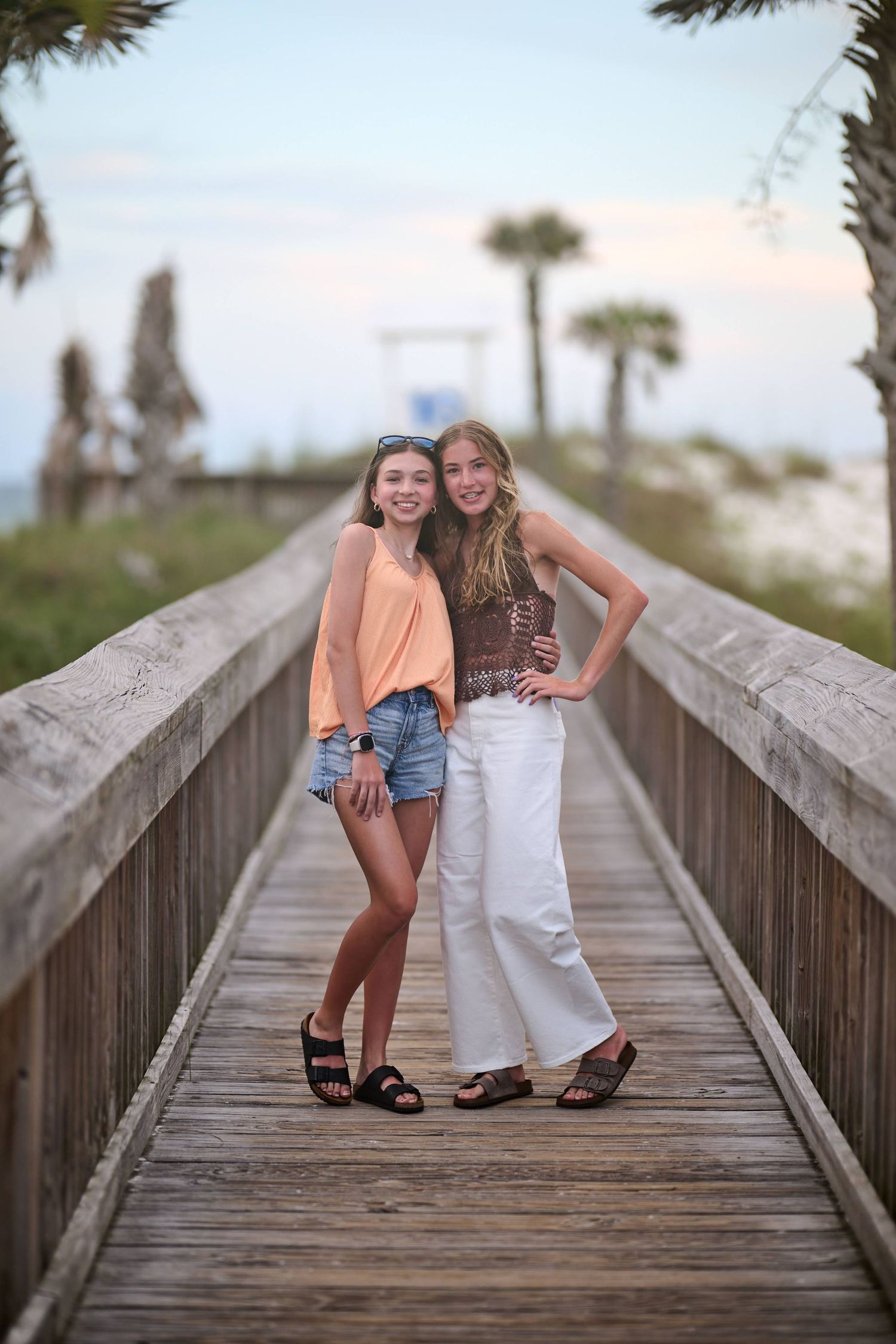 Two friends posing and laughing together on a wooden boardwalk with palm trees in the background at sunset.