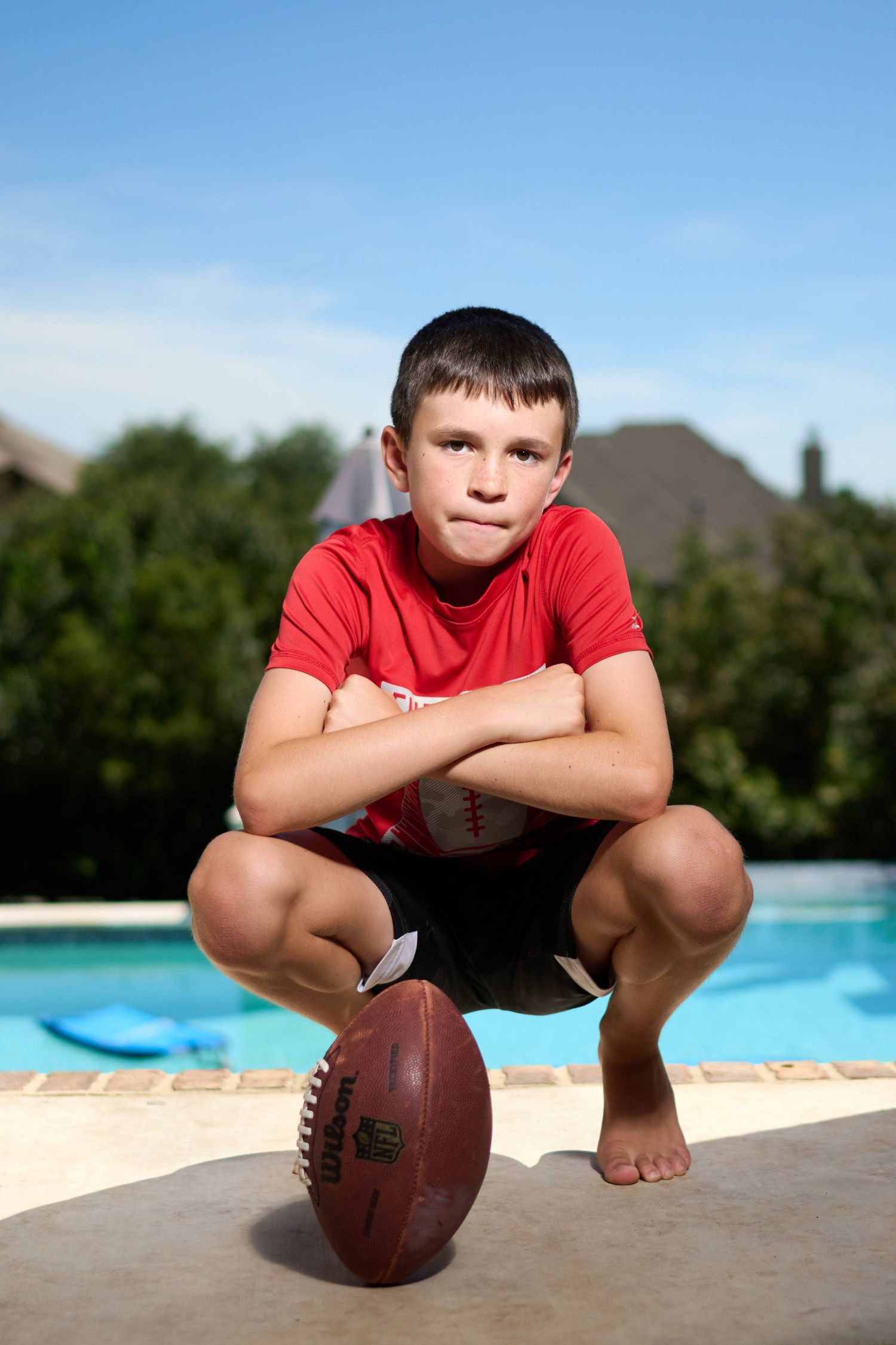 Young athlete crouching with football by pool edge wearing red shirt and black shorts.