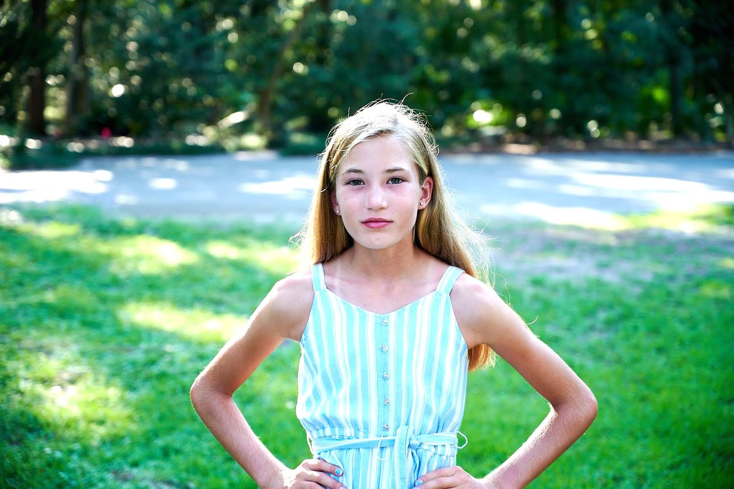 Portrait of a young person in a striped blue summer dress standing outdoors in a sunny park setting.