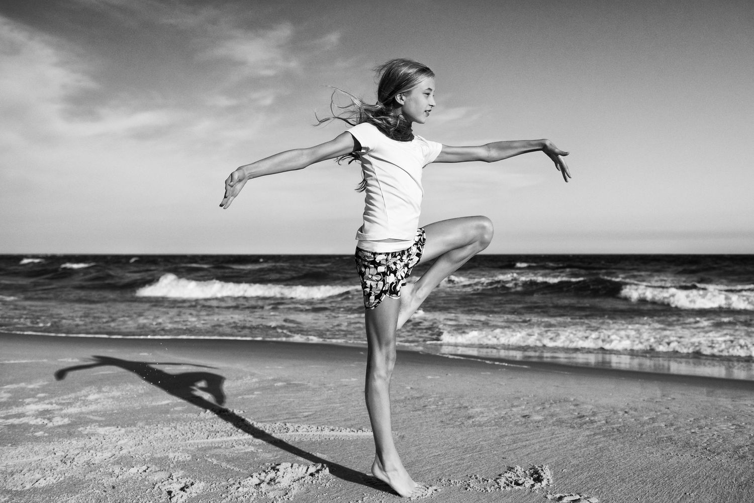 A graceful black and white photo of a dancer striking an arabesque pose on a beach with ocean waves in the background.