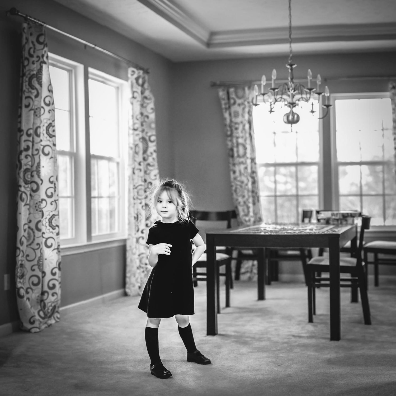 Black and white photo of small child in dress standing in elegant room with chandelier.