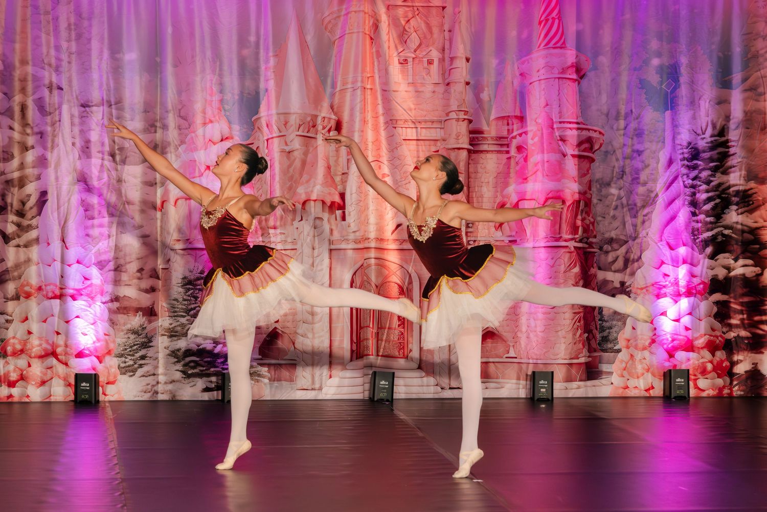 Two ballet dancers perform synchronized poses in burgundy tutus on stage with pink-lit winter scenery backdrop.