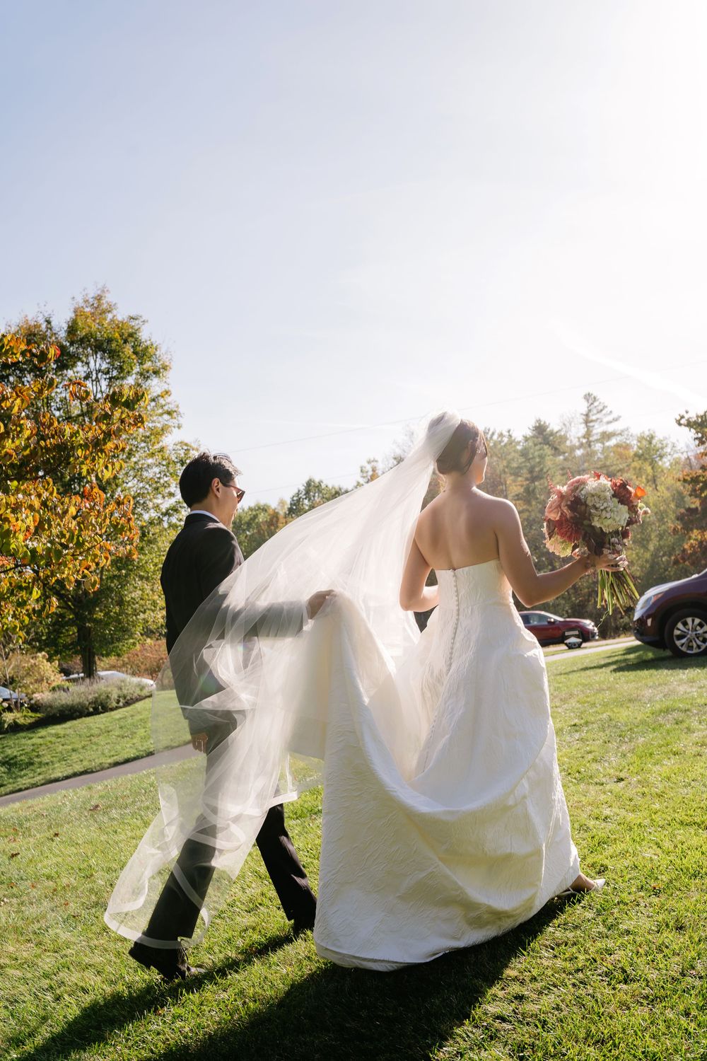 Bride and groom walking on green grass outdoors on sunny wedding day, veil flowing in breeze, bride holding bouquet.