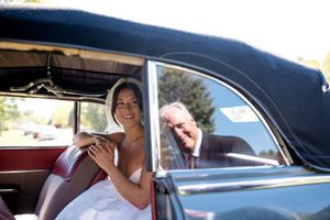 Bride in white dress smiles from vintage black car window with older gentleman beside her on wedding day.