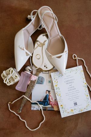 Elegant bridal flat lay with white heels, pearl necklace, gold jewelry, perfume, and floral wedding invitation on brown leather.