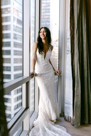 Bride in a white lace wedding gown with deep V-neckline stands by floor-to-ceiling windows with city skyline views.
