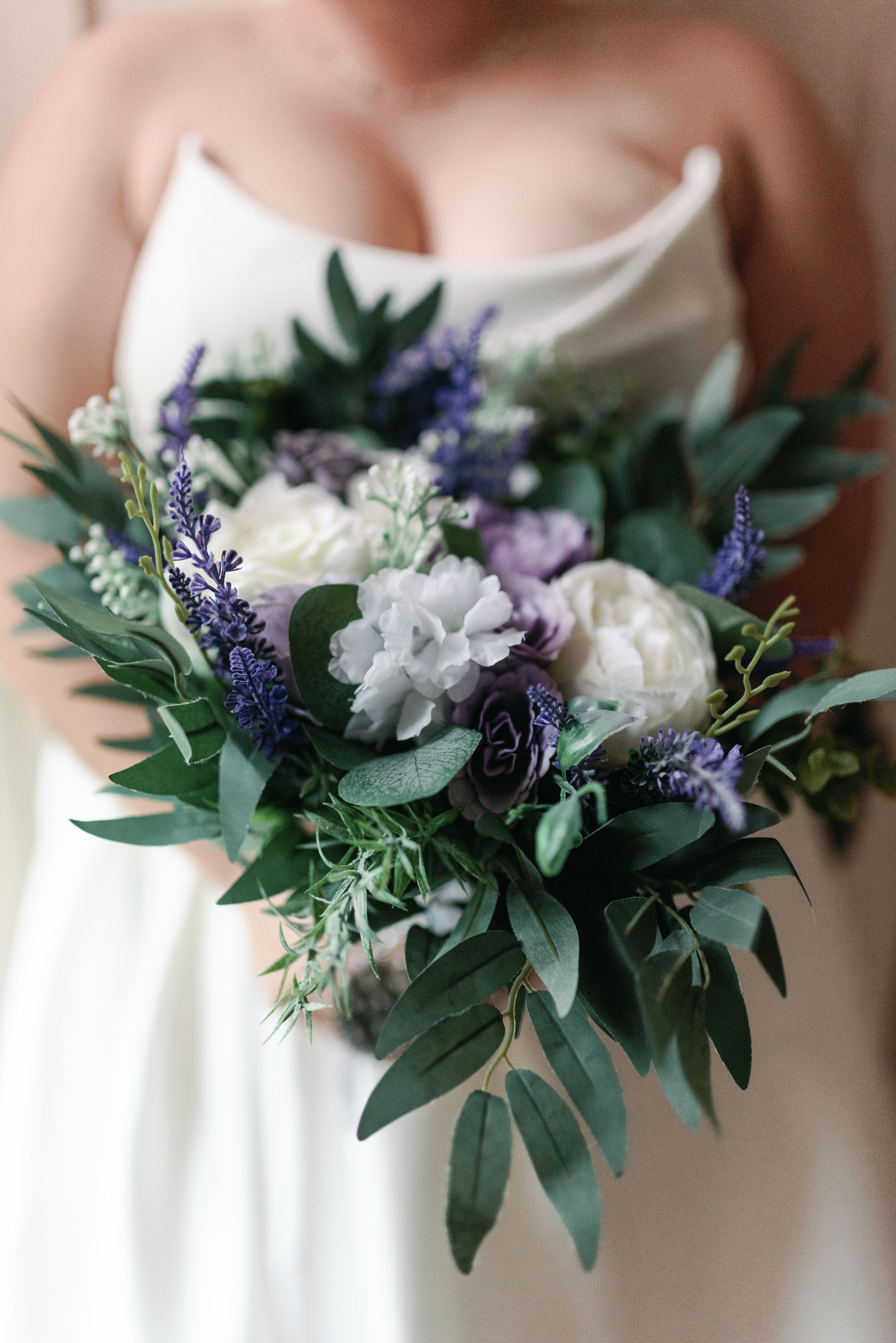 A romantic bridal bouquet with white roses, purple lavender sprigs, and eucalyptus leaves held against a white dress.