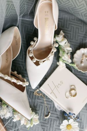 Elegant white bridal shoes with pearl details alongside wedding rings and floral accents.