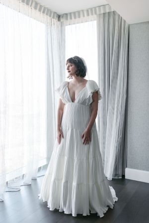 A flowing white wedding dress with empire waist and ruffled sleeves photographed against sheer curtains.