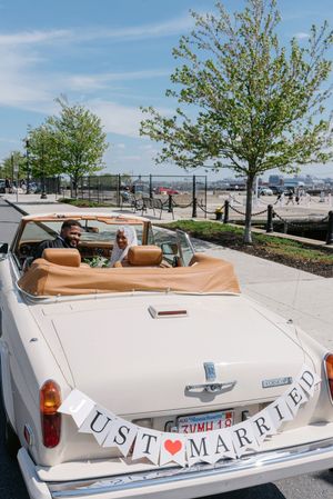 Newlyweds ride in a vintage white convertible car adorned with a Just Married banner on a sunny day.